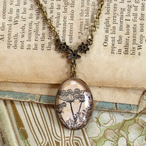 May include: A vintage-style necklace with a bronze chain and a glass pendant featuring a black and white illustration of a flower. The necklace is lying on a book with old, yellowed pages.