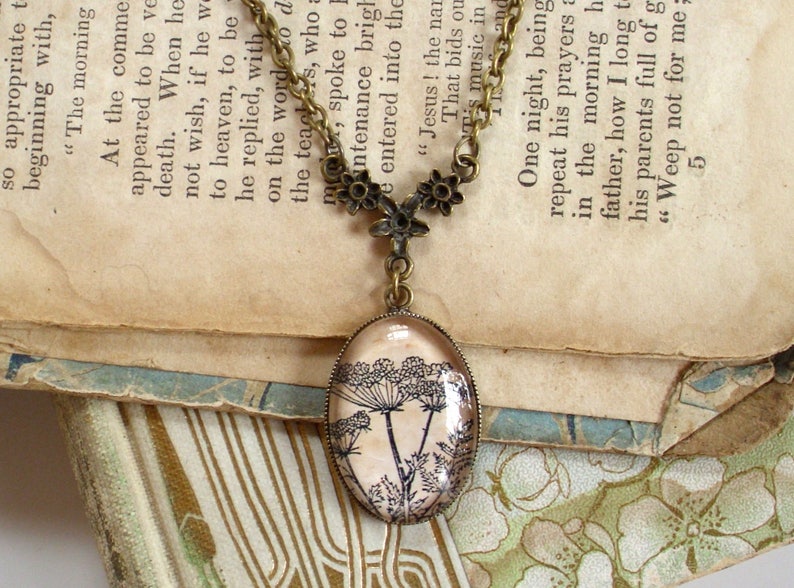 May include: A vintage-style necklace with a bronze chain and a glass pendant featuring a black and white illustration of a flower. The necklace is lying on a book with old, yellowed pages.