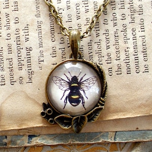 May include: A bronze necklace with a round glass pendant featuring a black and yellow bee illustration. The pendant is surrounded by a decorative bronze leaf design.