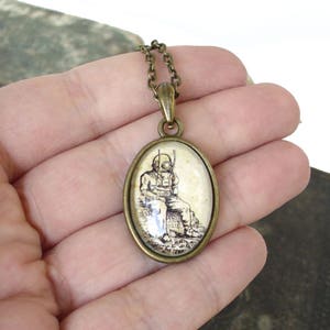 Op de afbeelding: Een bronzen ketting met een ovale hanger met een zwart-witte illustratie van een duiker met een helm en een speer. De hanger is in een glazen koepel gezet.