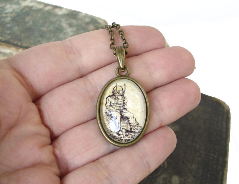 Op de afbeelding: Een bronzen ketting met een ovale hanger met een zwart-witte illustratie van een duiker met een helm en een speer. De hanger is in een glazen koepel gezet.
