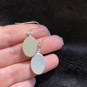 May include: A pair of silver oval-shaped earrings with a smooth, shiny finish. The earrings are hanging from a silver hook.