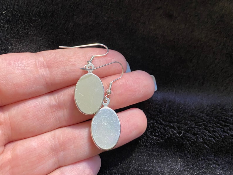 May include: A pair of silver oval-shaped earrings with a smooth, shiny finish. The earrings are hanging from a silver hook.