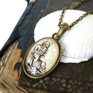 Op de afbeelding: Een bronzen ketting met een ronde glazen hanger met een vintage illustratie van een duiker met een helm. De hanger is geplaatst op een witte schelp.