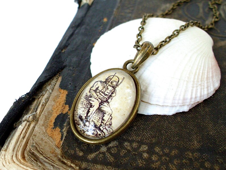 Op de afbeelding: Een bronzen ketting met een ronde glazen hanger met een vintage illustratie van een duiker met een helm. De hanger is geplaatst op een witte schelp.