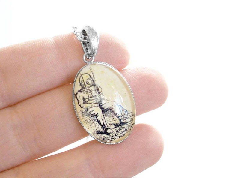 Op de afbeelding: Een zilveren ovale hangerketting met een vintage illustratie van een duiker in een duikpak. De illustratie is gedrukt op een cr&egrave;mekleurige achtergrond.