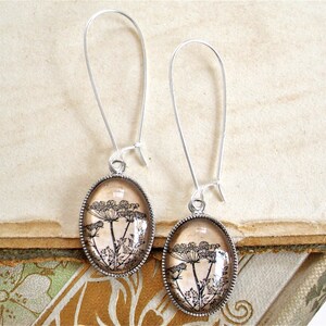 May include: A pair of silver dangle earrings with oval-shaped glass pendants featuring a black and white botanical print of a flower on a beige background.