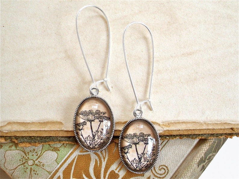May include: A pair of silver dangle earrings with oval-shaped glass pendants featuring a black and white botanical print of a flower on a beige background.