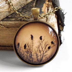 May include: A bronze necklace with a round glass pendant featuring a silhouette of a flock of crows perched on bare branches against a light brown background.