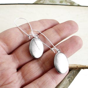 May include: A pair of silver dangle earrings with oval-shaped charms. The earrings are hanging from a person's hand.