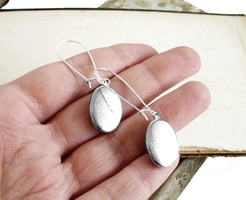 May include: A pair of silver dangle earrings with oval-shaped charms. The earrings are hanging from a person's hand.