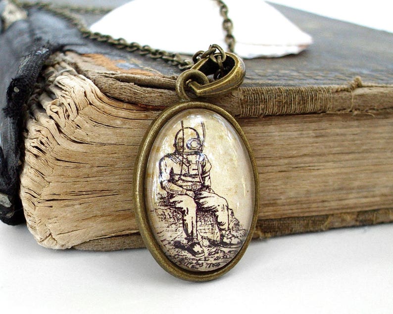 Op de afbeelding: Een bronzen ketting met een ovale glazen hanger met een vintage illustratie van een duiker in een duikpak. De hanger ligt op een oud lederen gebonden boek.