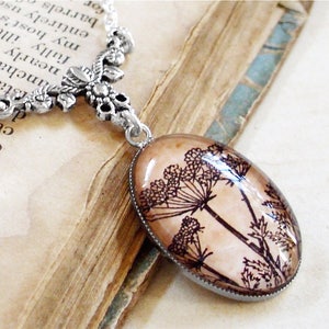 May include: A silver chain necklace with a delicate floral design pendant. The pendant features a glass oval with a pressed image of a brown and black wildflower.