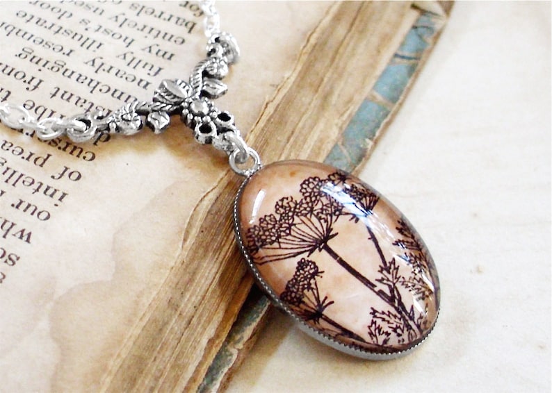 May include: A silver chain necklace with a delicate floral design pendant. The pendant features a glass oval with a pressed image of a brown and black wildflower.
