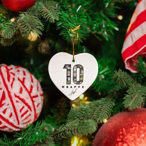A white heart-shaped ornament with the number 10 and the word "MBAPPED" is hanging on a Christmas tree, surrounded by ornaments and greenery.
