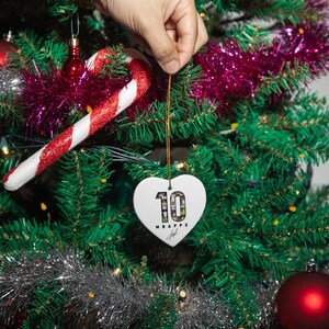 A hand is holding a heart-shaped ornament with the number 10 on it, hanging from a Christmas tree decorated with ornaments and tinsel.