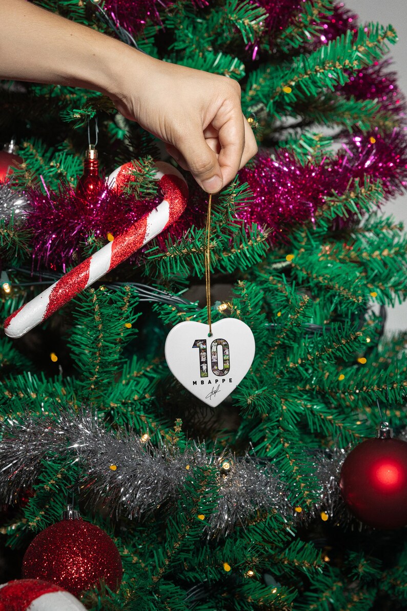A hand is holding a heart-shaped ornament with the number 10 on it, hanging from a Christmas tree decorated with ornaments and tinsel.