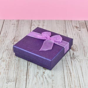 A purple gift box with a purple ribbon bow sits on a wooden surface against a pink background.