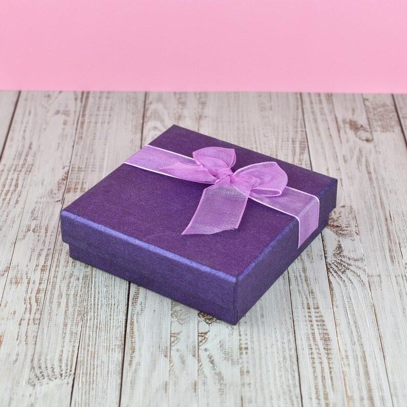 A purple gift box with a purple ribbon bow sits on a wooden surface against a pink background.