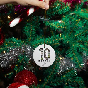 A hand is holding a white ornament with the number 10 on it, hanging from a Christmas tree decorated with red and white ornaments, tinsel, and other festive decorations.