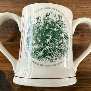 May include: Cream-colored ceramic mug with two handles. The mug features a green illustration of a group of people in military uniforms. The illustration is framed by a green circle. The mug has a purple stripe near the base.