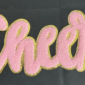 Op de afbeelding: Roze chenille patch met het woord "Cheer" in een sierlijk lettertype. De letters zijn omlijnd met gouden glitter, wat een contrast creëert. Geschikt voor kleding of accessoires.
