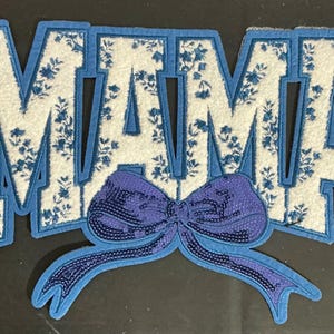 May include: A decorative patch with the word "MAMA" in large, block letters. The letters are white with a blue floral pattern and outlined in blue. A blue sequined bow is below the letters.