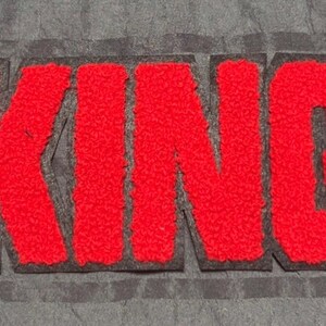 May include: A red chenille patch featuring the word KING in bold, block capital letters. The letters have a textured, fuzzy surface and are outlined with a thin black border, set against a dark gray fabric background with horizontal lines.