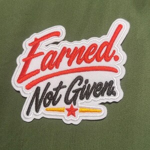 May include: A fabric patch featuring the red embroidered text Earned in a script font above the black embroidered text Not Given in a cursive font, with a small red star and two yellow horizontal lines centered below on a white background.
