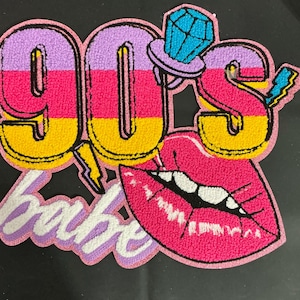 May include: A colorful patch with the text "90's babe" in a retro design. The patch features a large pink lip, a diamond ring, and a lightning bolt. The numbers "90" are in yellow, pink, and purple stripes.