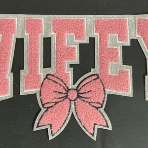 May include: A pink and silver embroidered patch with the word "WIFEY" in large, block letters. A pink bow is below the text. The patch is on a black background.