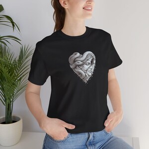 Könnte beinhalten: Ein schwarzes kurzärmeliges T-Shirt mit einem zentralen Grafikdruck eines menschlichen Gesichts, das von einer Schlange umschlungen ist und eine Herzform bildet. Die Illustration ist in Grau- und Weißtönen mit detaillierten Linien gehalten.