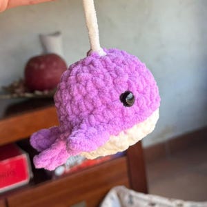 Puede incluir: Un juguete de ballena hecho a mano, de ganchillo, en tonos morados y blanco. La ballena tiene un ojo negro y una parte inferior blanca. Suspendido por un limpiapipas blanco. Juguete suave y texturizado.