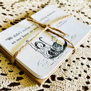 May include: A stack of cards with a black and white illustration of a girl and a bear on a boat. The cards are tied together with twine and have the quote "We didn't realize we were making memories, we just knew we were having fun." by A.A. Milne, Winnie the Pooh.