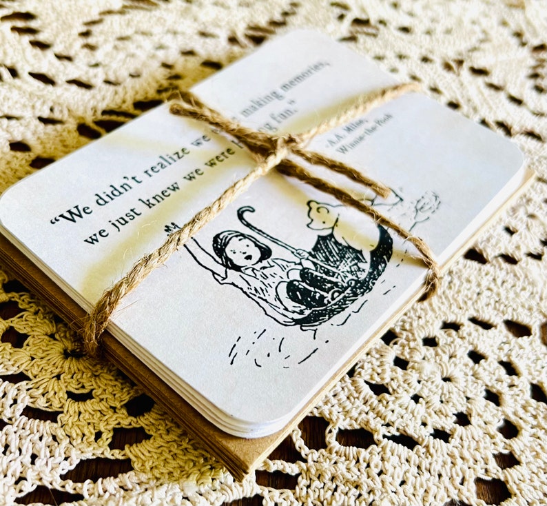 May include: A stack of cards with a black and white illustration of a girl and a bear on a boat. The cards are tied together with twine and have the quote "We didn't realize we were making memories, we just knew we were having fun." by A.A. Milne, Winnie the Pooh.
