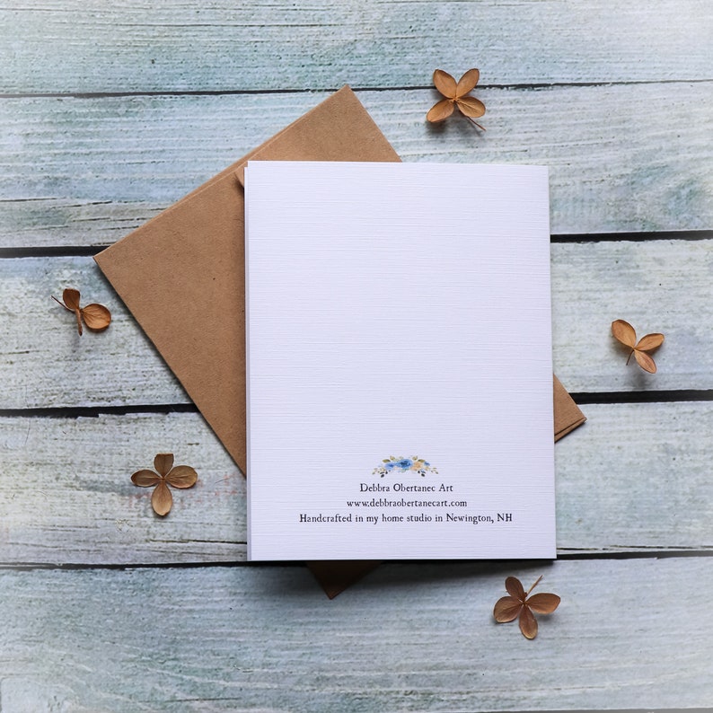 May include: A blank white card with lined paper and a blue floral design at the bottom. The card has the text "Debra Obertace Art" and "www.debraobertaceart.com" and "Handcrafted in my home studio in Newington, NH". The card is on a light blue wooden surface with brown dried flowers.