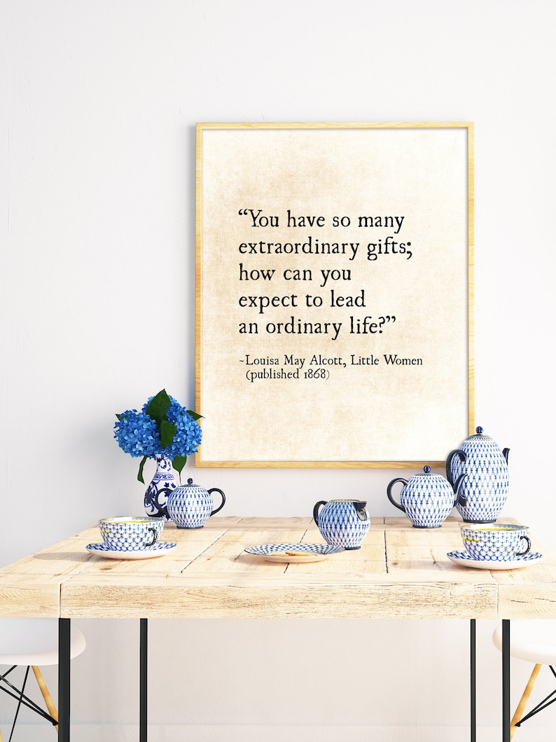 May include: A framed print with a quote from Louisa May Alcott's "Little Women" that reads, "You have so many extraordinary gifts; how can you expect to lead an ordinary life?" The quote is printed in black text on a cream background. The print is sitting on a wooden table with blue and white teacups and a bouquet of blue hydrangeas.