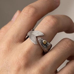 Op de afbeelding: Een zilverkleurige ring met een visontwerp. De ring wikkelt zich om de vinger, met een vissenstaart aan de ene kant en een vissenkop met een goudkleurig oog aan de andere kant. De ring heeft een geoxideerde afwerking.