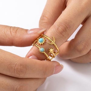 Può includere: Anello color oro con disegni celesti. L'anello presenta un motivo a sole, luna e stella, con piccole pietre color turchese. L'anello è indossato su un dito, mostrando i suoi dettagli e il suo design intricati.