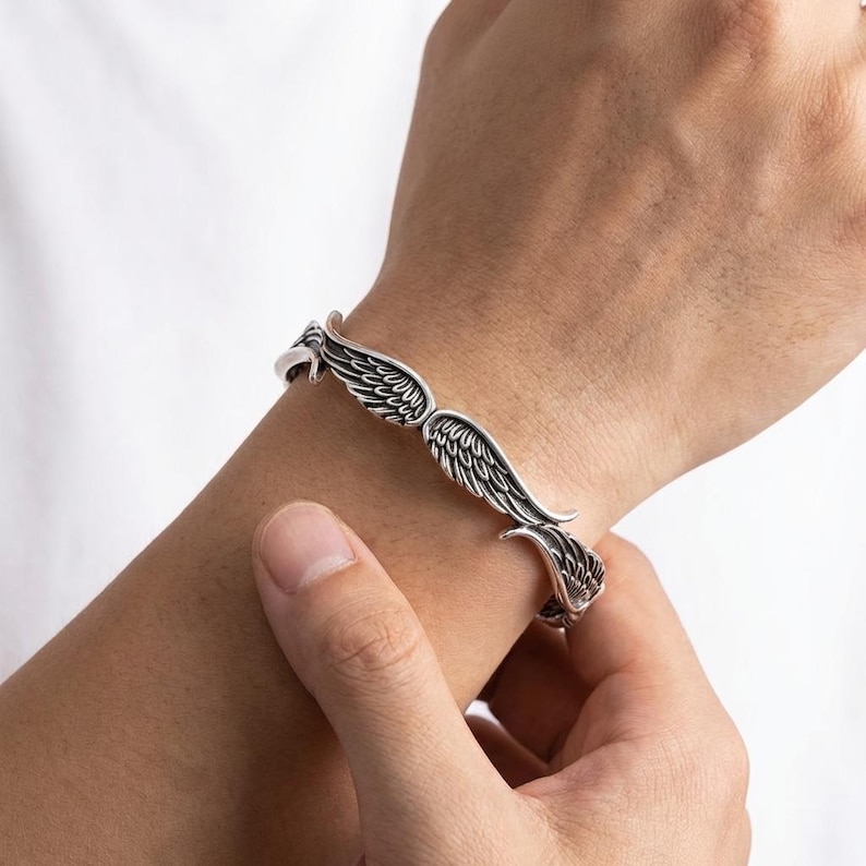 K&ouml;nnte beinhalten: Ein silberfarbenes Armband mit einem Fl&uuml;geldesign. Das Armband, das am Handgelenk getragen wird, hat detaillierte Federmuster. Die Fl&uuml;gel umschlie&szlig;en das Handgelenk und schaffen ein einzigartiges und stilvolles Accessoire.