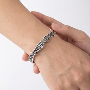 K&ouml;nnte beinhalten: Ein silberfarbenes Armband mit Fl&uuml;geldesign. Das Armband hat detaillierte Federmuster und schmiegt sich um das Handgelenk. Die Fl&uuml;gel sind so positioniert, dass sie das Handgelenk zu umh&uuml;llen scheinen.