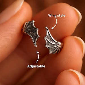Puede incluir: Un par de puños de oreja ajustables con forma de ala de color plateado. Los puños de oreja están diseñados para parecerse a alas de murciélago. La imagen muestra los puños de oreja en una vista de cerca, con un puño separado del otro.