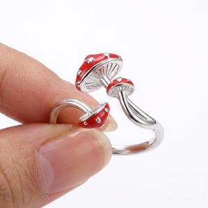 May include: A silver ring with a mushroom design. The ring features three mushrooms with red caps and white spots. The underside of the mushroom caps are white with detailed gills. The ring is held by a person's fingers.