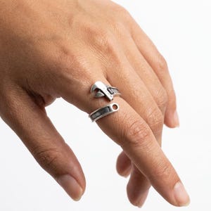 Puede incluir: Un conjunto de anillos plateados con un diseño de llave inglesa. El anillo está dividido en dos partes, una que se asemeja a la cabeza de la llave y otra al mango. La cabeza de la llave está colocada en el dedo índice y el mango en el dedo corazón.