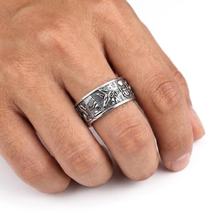 Peut inclure: Une bague argentée avec un motif gravé de hiéroglyphes égyptiens. La bague large présente divers symboles et figures, notamment des scarabées et d'autres motifs de l'Égypte ancienne. La bague est portée au doigt.