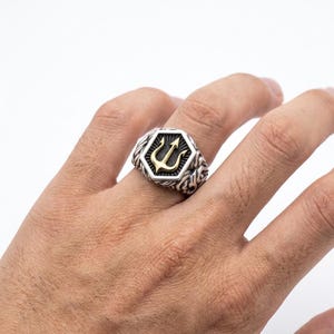 Puede incluir: Un anillo plateado con un emblema de tridente dorado en un marco hexagonal. El anillo tiene una banda detallada y texturizada. El tridente, una lanza de tres puntas, es un símbolo de poder. El anillo se lleva en un dedo.