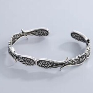 K&ouml;nnte beinhalten: Ein silberfarbenes Armreif-Armband mit detailliertem Fl&uuml;geldesign. Das offene Armband verf&uuml;gt &uuml;ber mehrere S&auml;tze stilisierter Fl&uuml;gel, die ein dekoratives und symbolisches Accessoire schaffen. Das Armband ist f&uuml;r das Handgelenk konzipiert.