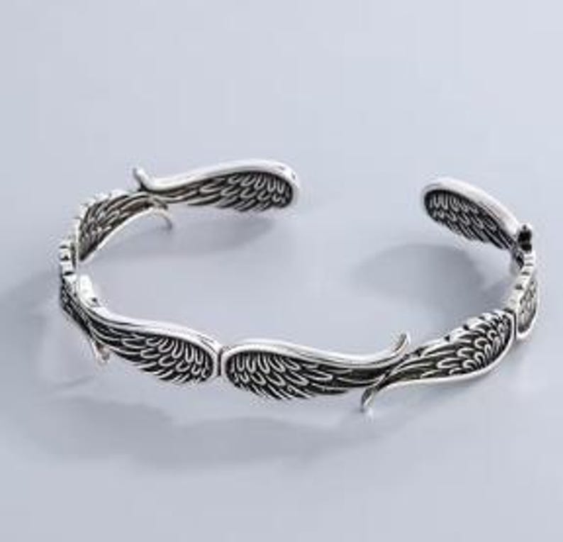 K&ouml;nnte beinhalten: Ein silberfarbenes Armreif-Armband mit detailliertem Fl&uuml;geldesign. Das offene Armband verf&uuml;gt &uuml;ber mehrere S&auml;tze stilisierter Fl&uuml;gel, die ein dekoratives und symbolisches Accessoire schaffen. Das Armband ist f&uuml;r das Handgelenk konzipiert.