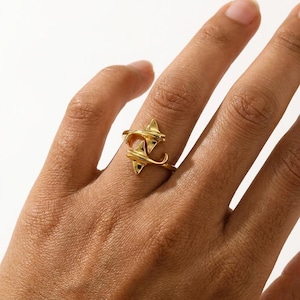 Op de afbeelding: Een goudkleurige ring met een gestileerd ontwerp van twee manta's die tegenover elkaar staan. De ring heeft een gladde, gepolijste afwerking en wordt op een vinger gedragen. Het ontwerp bevat driehoekige en gebogen elementen.
