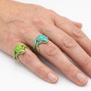 Puede incluir: Dos anillos con forma de rana, uno verde y otro turquesa, se llevan en los dedos. Los anillos tienen detalles de rana y están hechos de un material brillante. Los anillos son un accesorio único.